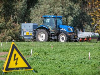 Blue tractor in a weedy riverside paddock with a front end brace on it with flaps.