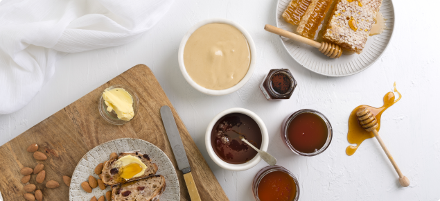 Display of honey and honeycomb on a white plate, bread, almonds and butter on a cutting board and different jams
