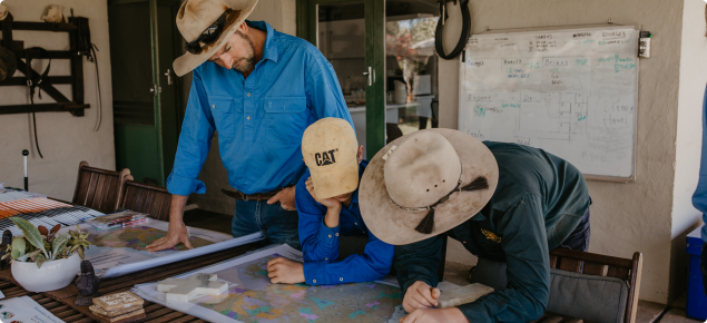 A man, woman and child dressed in pastoral attire look over plans on a table