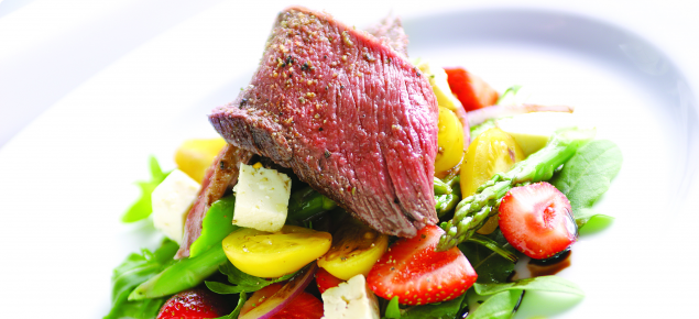 A salad, including cheese and strawberries, topped with a slice of meat on a white serving platter.