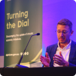 Adam Carrel seated on stage with a Turning the Dial banner in the background
